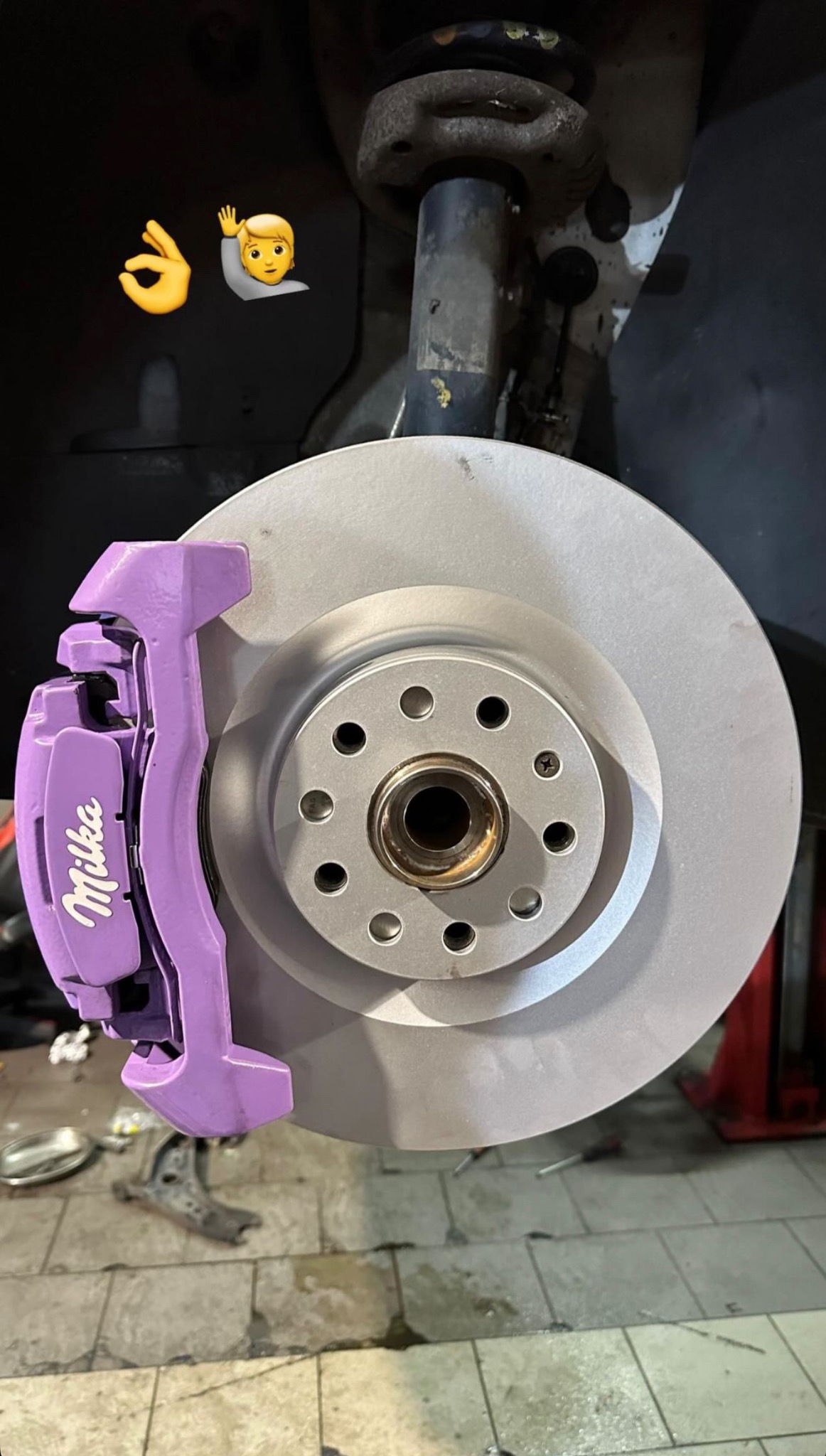 Tarcza hamulcowa z fioletowym zaciskiem marki Milka, zamontowana w samochodzie. Widoczne emoji. Nietypowy kolor zacisku wyróżnia to ujęcie.