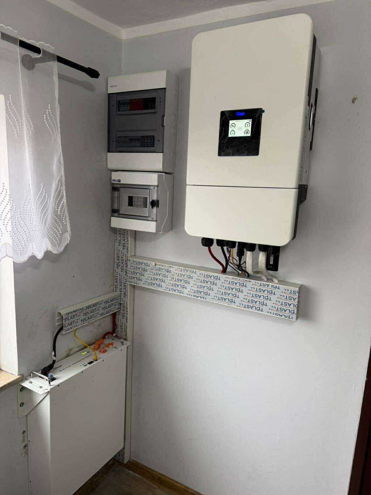 Biały inwerter fotowoltaiczny i akumulator energii zamontowane na ścianie wewnątrz budynku, widoczne kable i elementy instalacji elektrycznej.