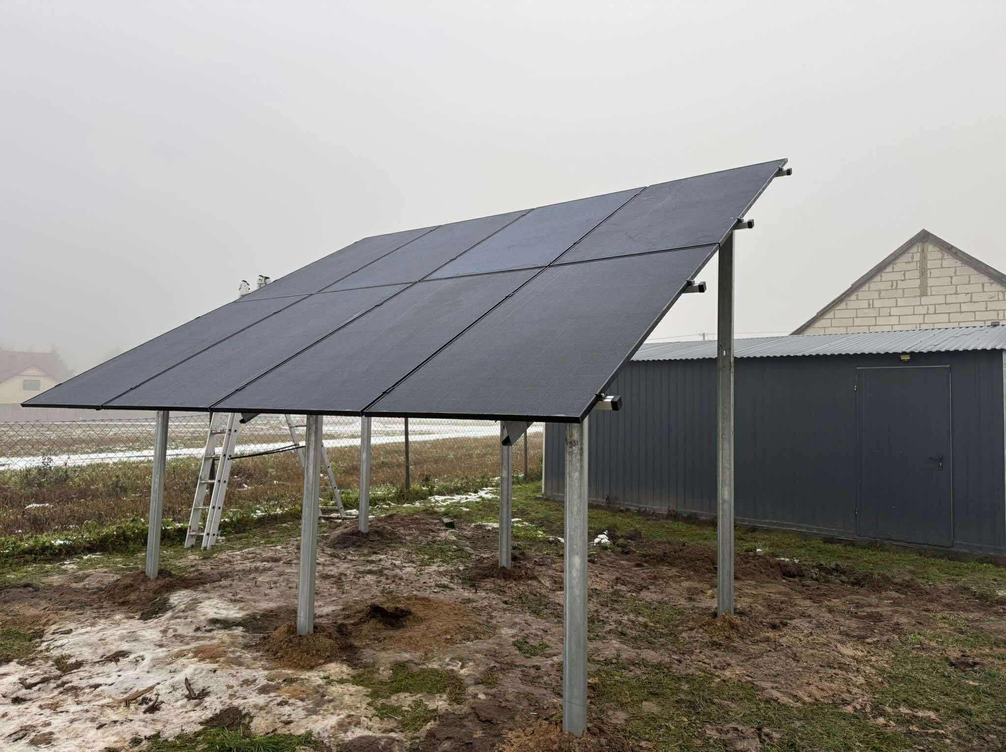 Montaż paneli fotowoltaicznych na konstrukcji wsporczej w Spławiu. Widoczna drabina i przygotowany teren pod instalację. Pochmurne niebo w tle.