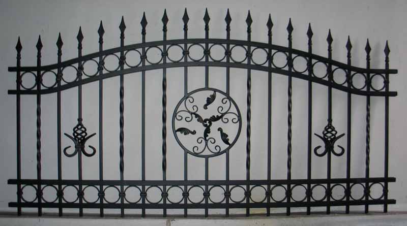 Fragment czarnego, kutego ogrodzenia z dekoracyjnymi elementami: kolcami, okręgami, spiralnymi prętami i rozetą z motywem roślinnym na szarym tle.