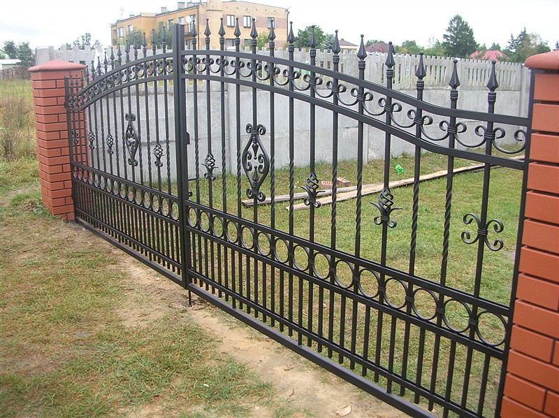 Czarna, dwuskrzydłowa brama wjazdowa z kutymi zdobieniami, osadzona na ceglanych słupkach, na tle trawnika i betonowego ogrodzenia.