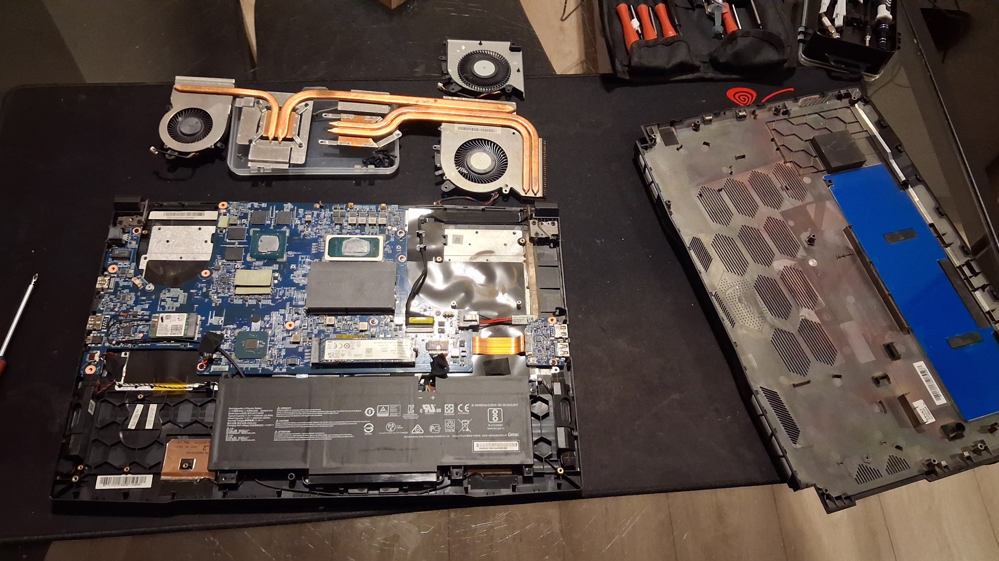 Rozłożony laptop z widoczną płytą główną, systemem chłodzenia z miedzianymi rurkami cieplnymi i wentylatorami. Obok leży dolna obudowa i narzędzia.