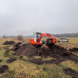 KOLD-BAU budownictwo i nieruchomości - Pomarańczowa mini koparka na polu, wykop pod rury kanalizacyjne, widoczne ślady opon i wykopana ziemia. W tle drzewa i pochmurne niebo.