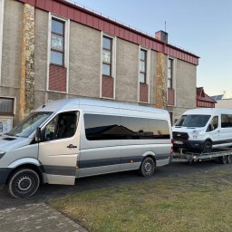 KOLD-BAU budownictwo i nieruchomości - Srebrny bus Mercedes z otwartymi drzwiami i biały Ford na lawecie przed budynkiem z oknami witrażowymi. Transport pojazdów, widok z boku, wczesny wieczór.