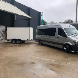 KOLD-BAU budownictwo i nieruchomości - Srebrny bus Mercedes z białą przyczepą na zewnątrz budynku przemysłowego. Na ścianie budynku drabina. Mokra nawierzchnia placu.