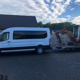 KOLD-BAU budownictwo i nieruchomości - Biały bus z przyczepą transportuje pomarańczową mini-koparkę na tle brukowanej nawierzchni i domu z ciemnym dachem, pod błękitnym niebem z chmurami.