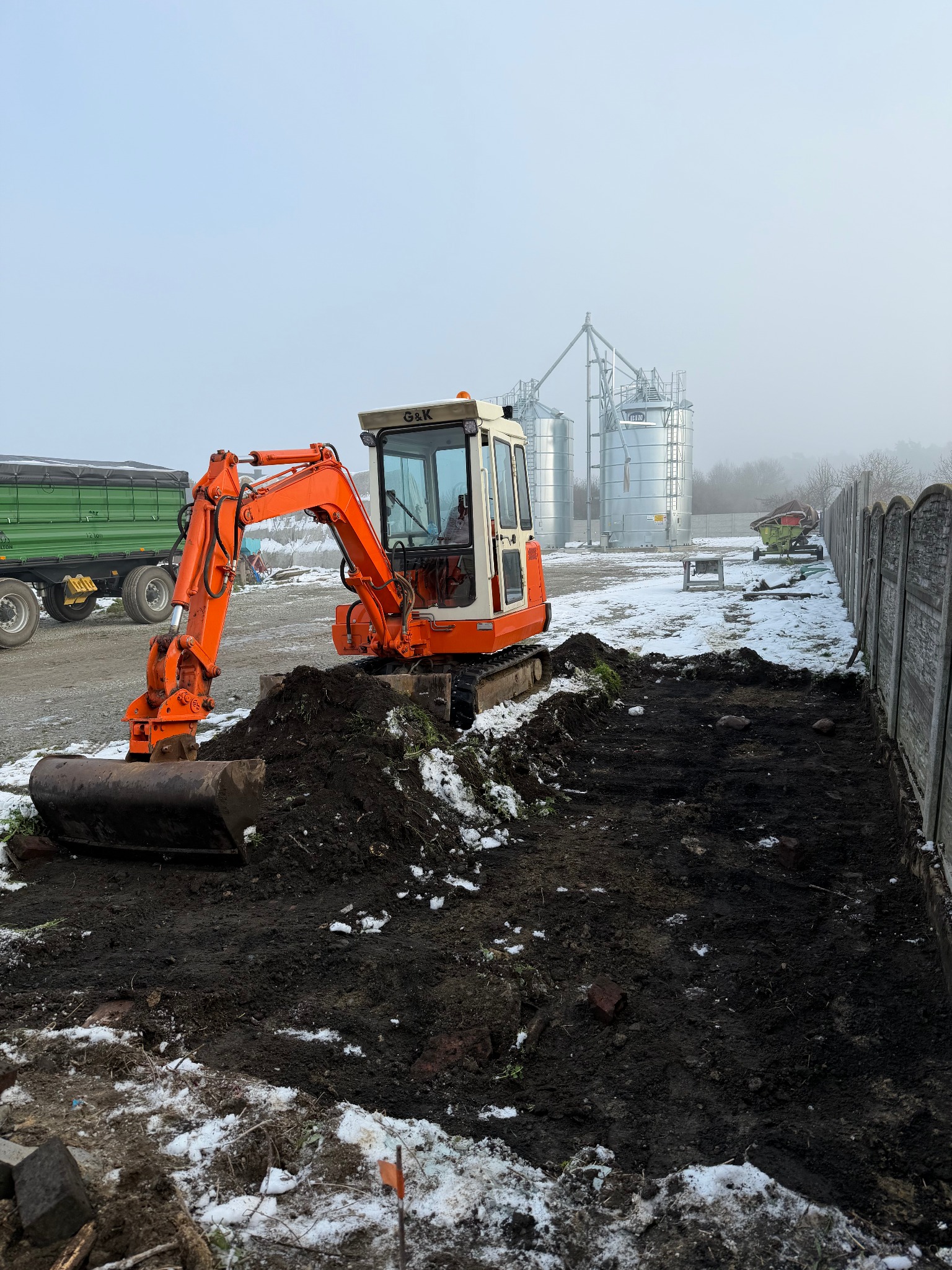 Pomarańczowa mini koparka G&K w trakcie prac ziemnych przy ogrodzeniu, z widocznymi silosami w tle i resztkami śniegu na ziemi.