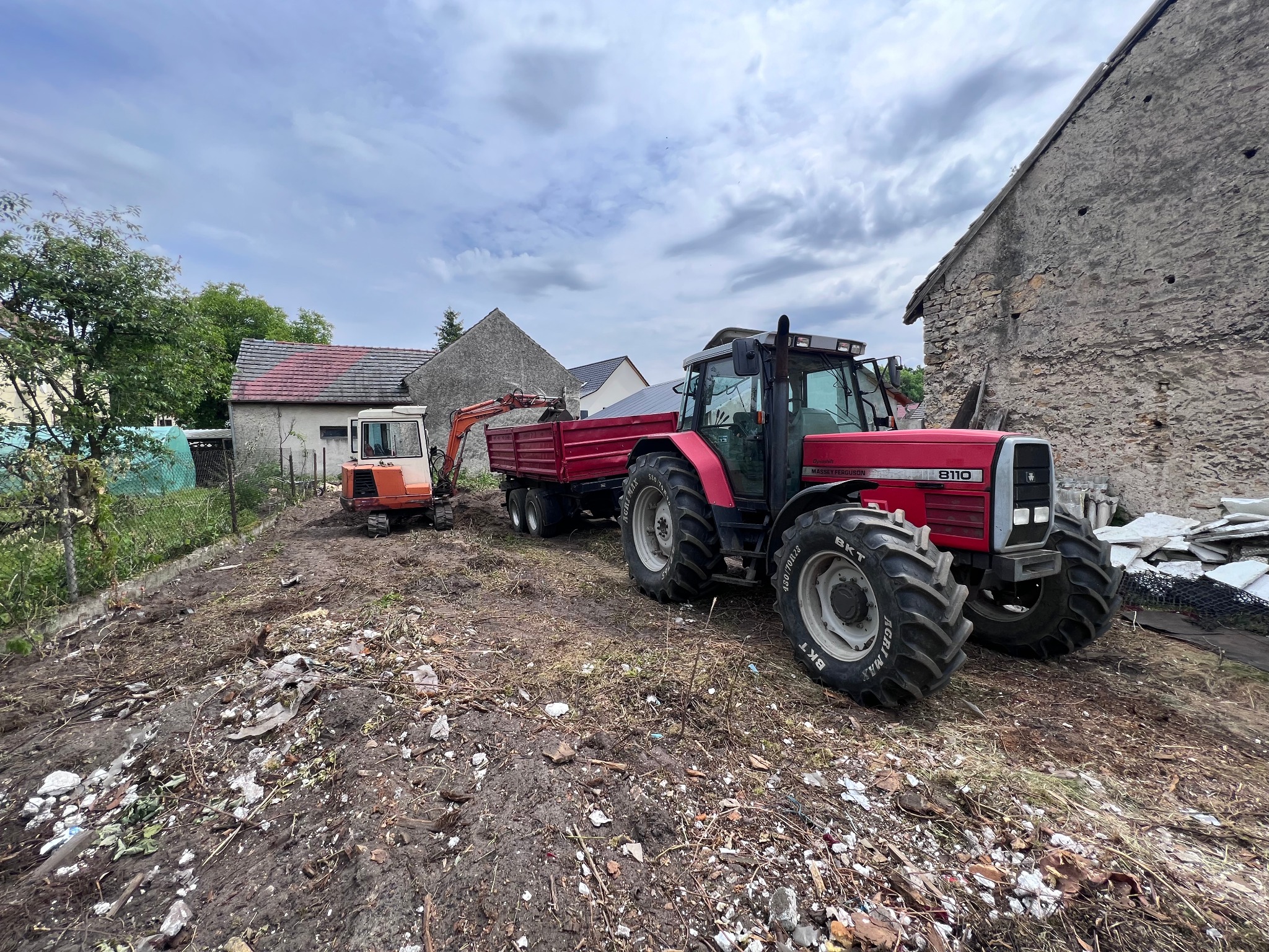 Czerwony traktor Massey Ferguson 8110 z przyczepą i mała koparka na placu budowy, w tle budynki. Prace ziemne w toku, widoczne ślady po rozbiórce.