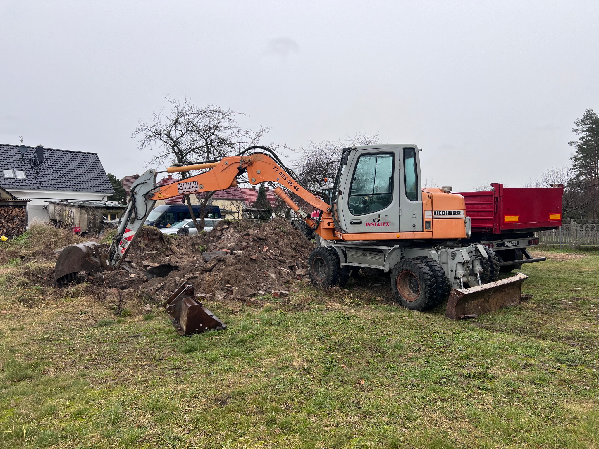 Pomarańczowo-szara koparka Liebherr na trawiastym terenie z wykopaną ziemią i dodatkową łyżką obok. W tle dom z ciemnym dachem i drzewa bez liści.