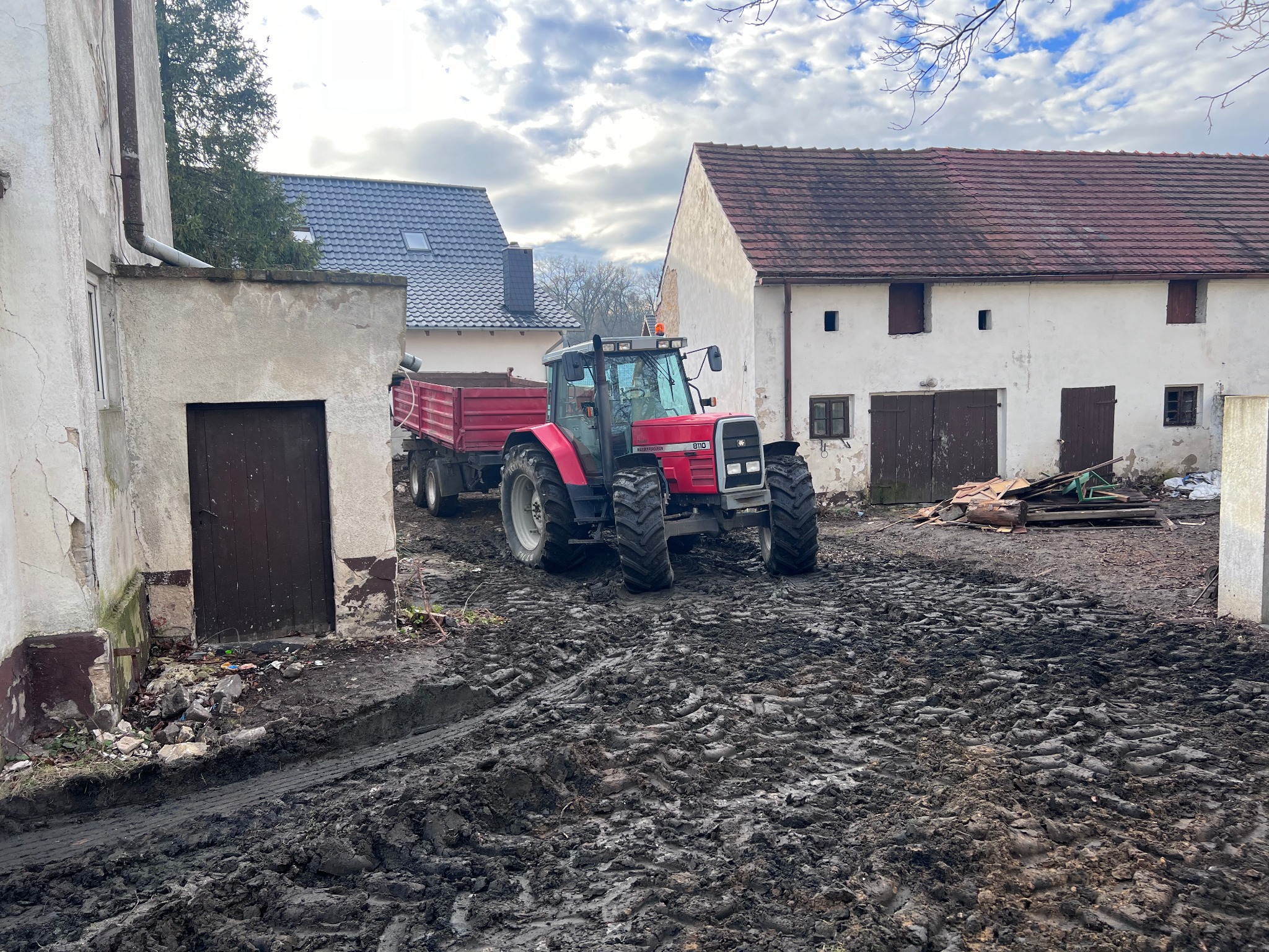 Czerwony traktor z przyczepą na błotnistym terenie budowy, ślady opon, budynki w tle, pochmurne niebo. Widoczne ślady prac ziemnych.