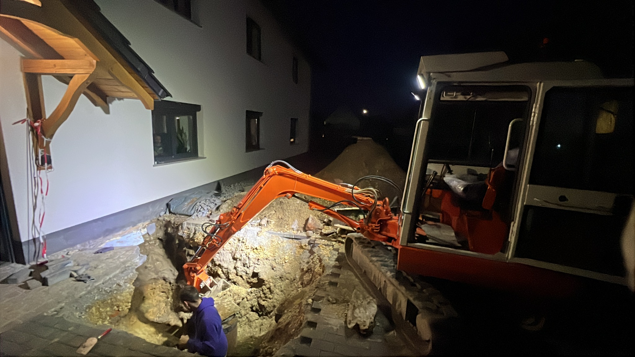 Pomarańczowa mini koparka w nocnej scenerii, wykonująca wykop w pobliżu budynku. Widoczny pracownik w dole wykopu i sterta ziemi w tle.
