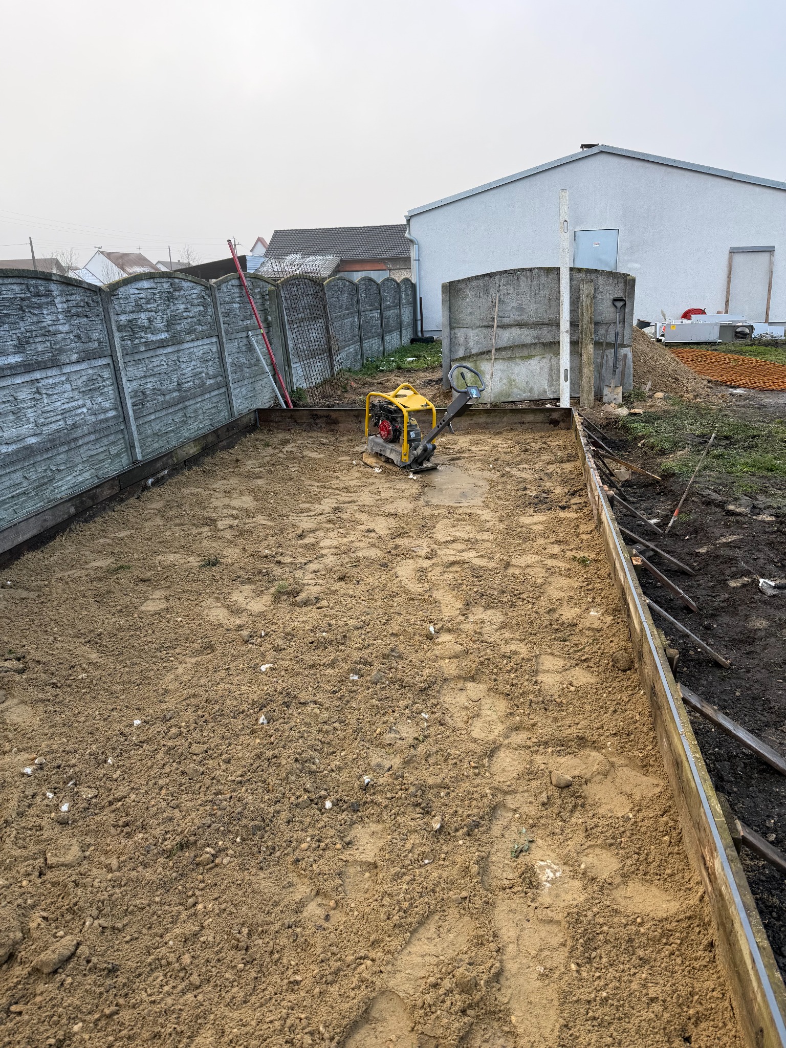 Przygotowany teren pod budowę z żółtą zagęszczarką w tle, otoczony drewnianym szalunkiem i betonowym ogrodzeniem. Widoczny budynek w oddali.