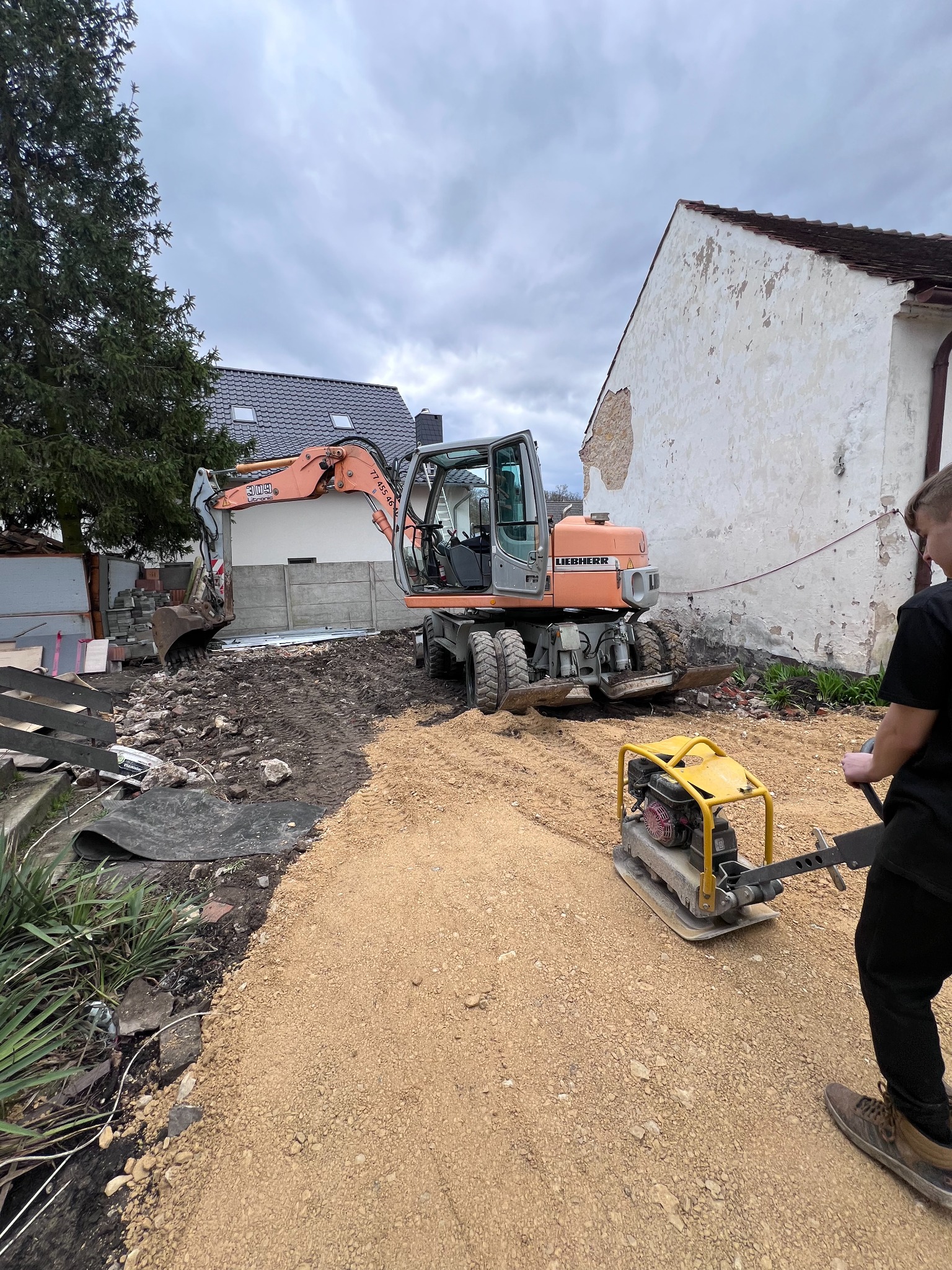 Pomarańczowa koparka Liebherr i zagęszczarka na piasku, przygotowanie terenu pod budowę, widoczny fragment budynku z uszkodzonym tynkiem i osoba obsługująca zagęszczarkę.