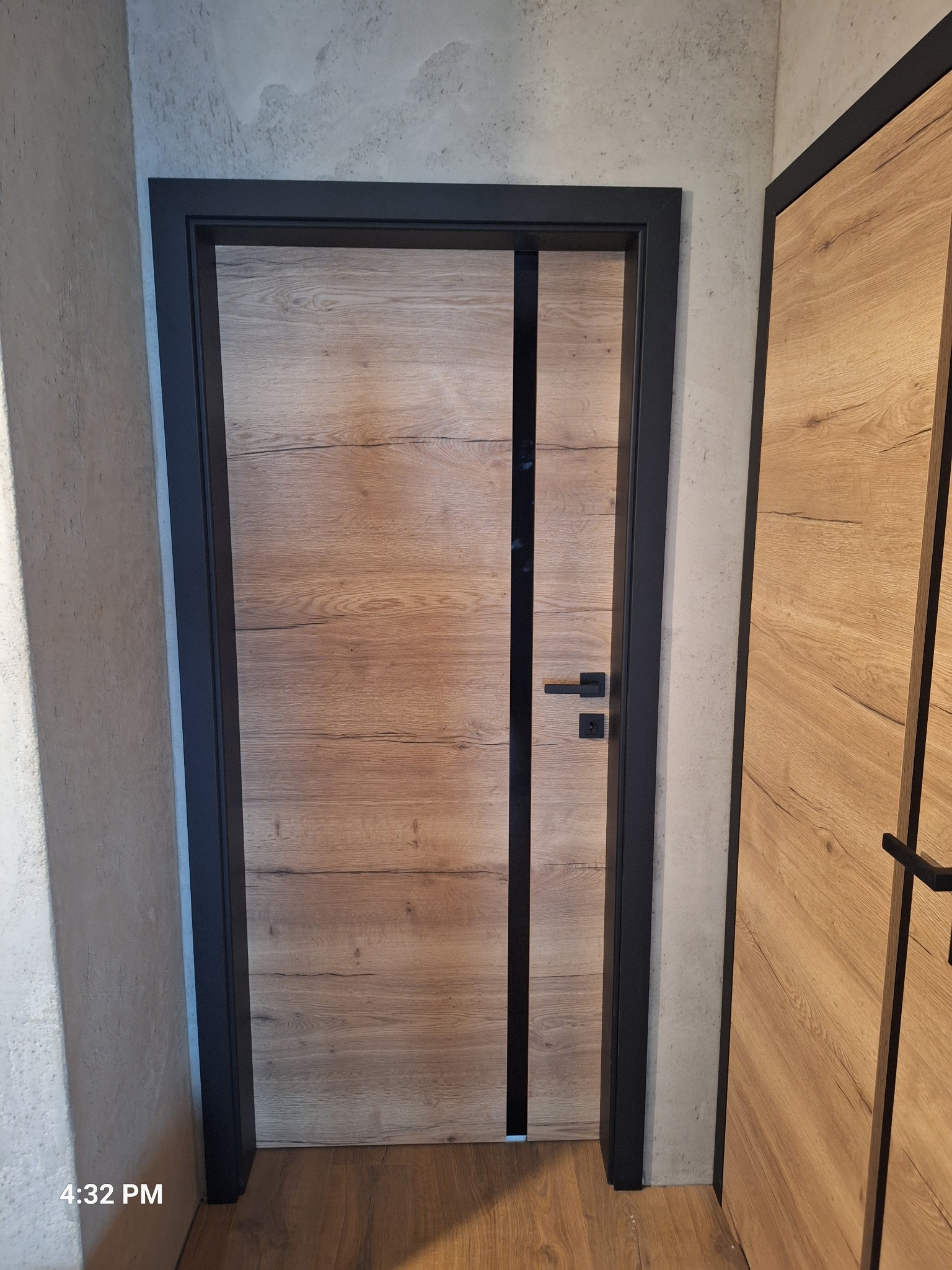 Zamknięte drzwi wewnętrzne z drewnopodobnym panelem i czarną ramą, widoczne na tle szarej ściany. Obok fragment drugich drzwi z podobnym wykończeniem.