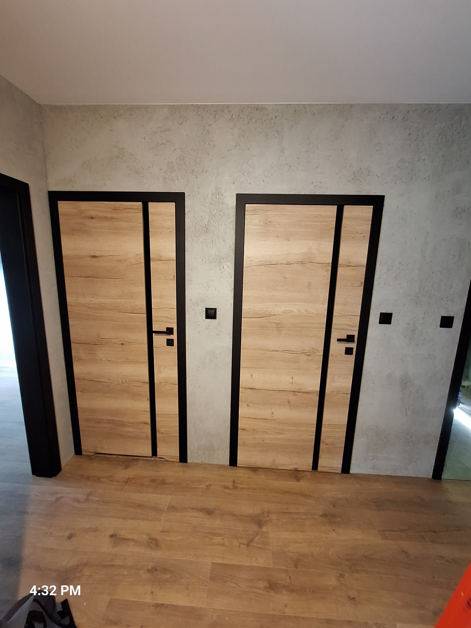 Dwa zamontowane, drewnopodobne drzwi z czarnymi ościeżnicami na tle szarej ściany o fakturze betonu i drewnianej podłogi. Widoczne czarne klamki i włączniki.