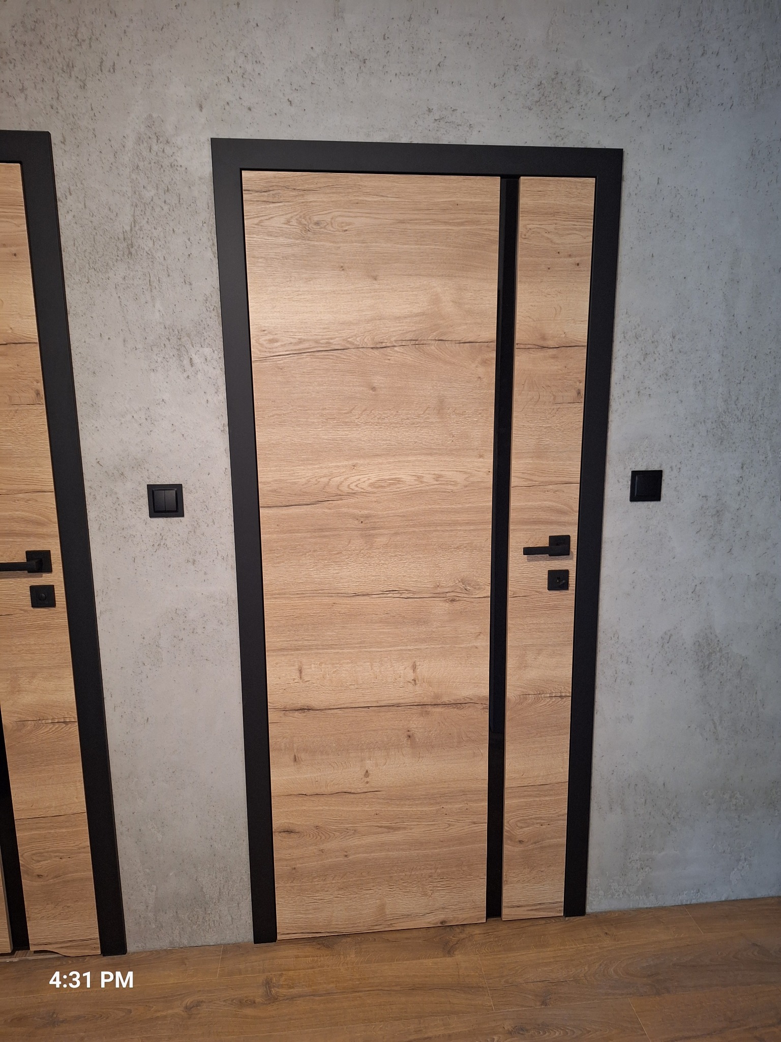 Dwa nowoczesne, drewnopodobne drzwi z czarnymi ramami na tle szarej ściany. Widoczne detale klamek i włączników. Podłoga drewniana.