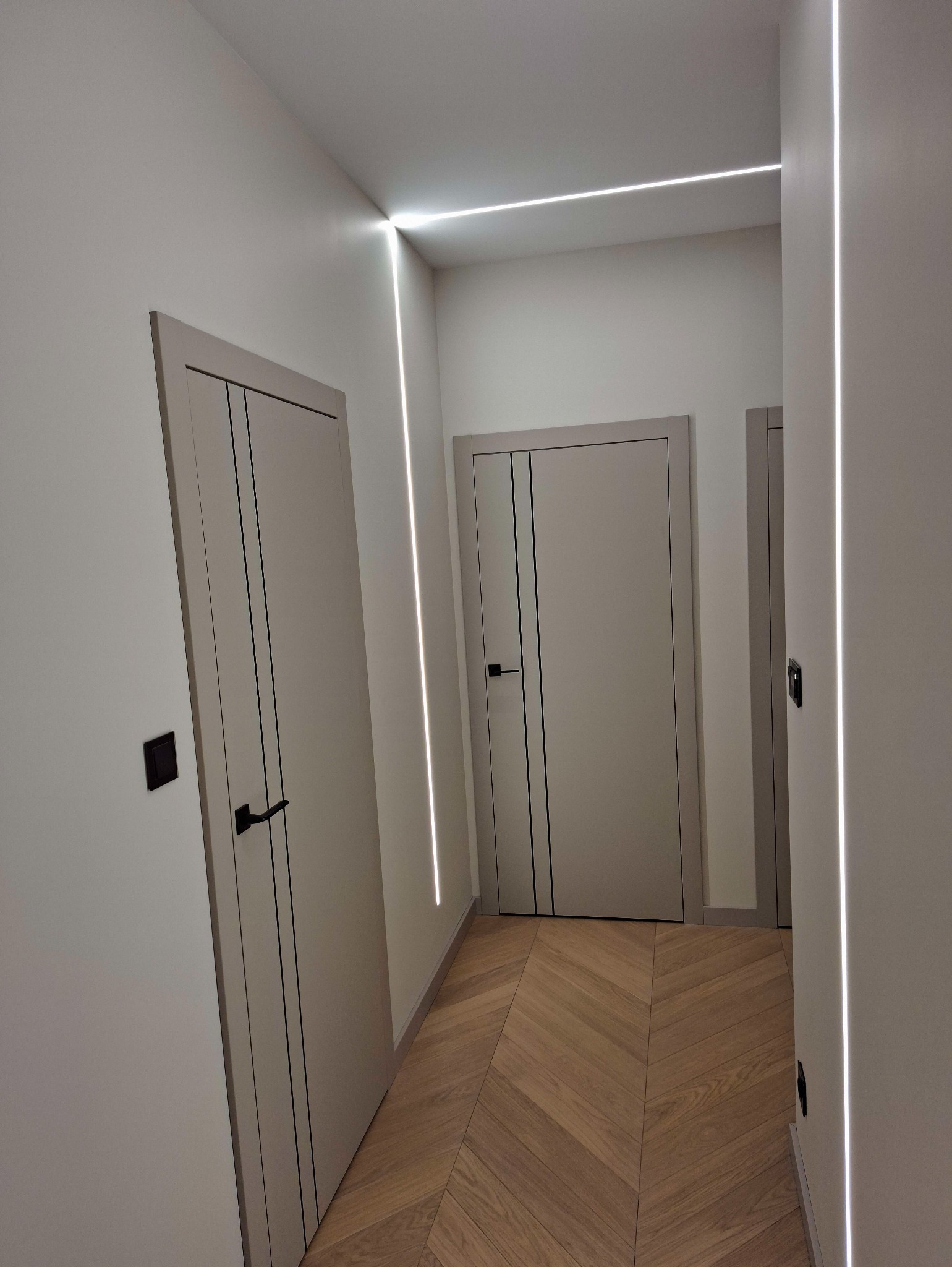 Nowoczesny korytarz z trzema szarymi drzwiami z czarnymi akcentami i parkietem w jodełkę. Oświetlenie LED wzdłuż ścian tworzy minimalistyczny efekt.