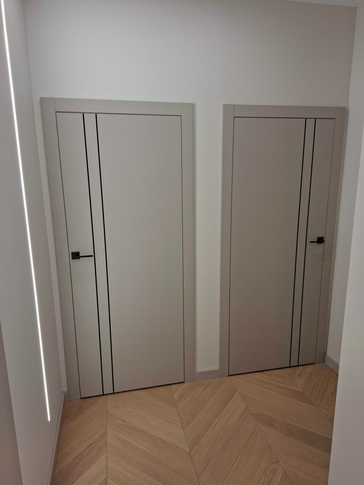 Dwa szare, nowoczesne drzwi wewnętrzne z czarnymi akcentami i klamkami, zamontowane w jasnym pomieszczeniu z parkietem ułożonym we wzór jodełki.