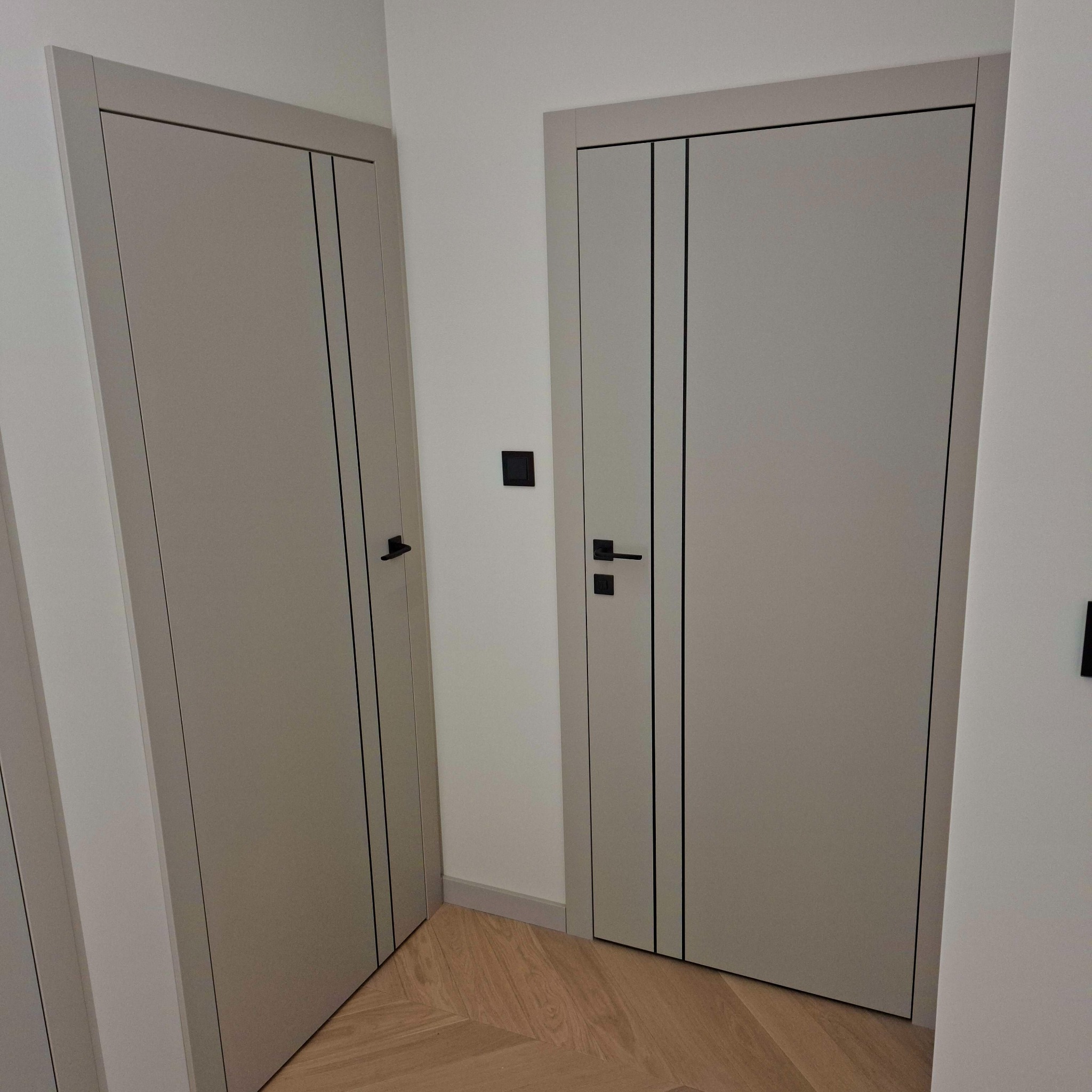 Dwa beżowe, nowoczesne drzwi wewnętrzne z czarnymi akcentami, zamontowane w białym korytarzu z drewnianą podłogą. Minimalistyczny design.