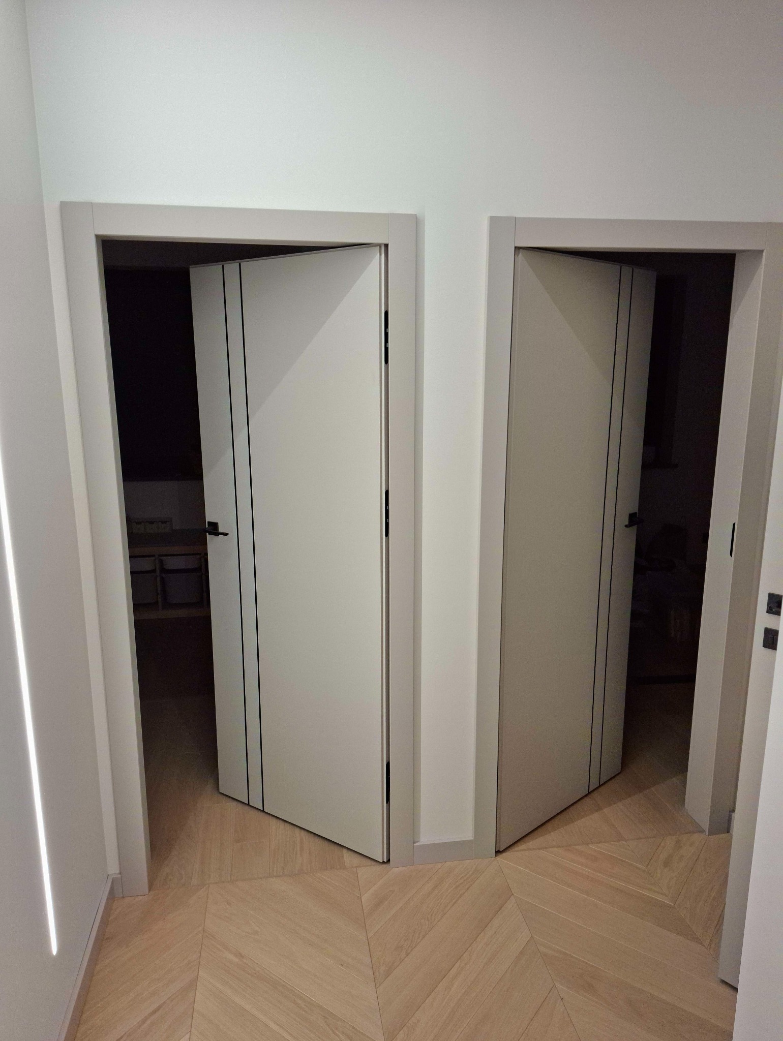 Dwa białe, nowoczesne drzwi wewnętrzne z czarnymi paskami, zamontowane w jasnym pomieszczeniu z parkietem ułożonym we wzór jodełki. Jedne drzwi otwarte, drugie zamknięte.