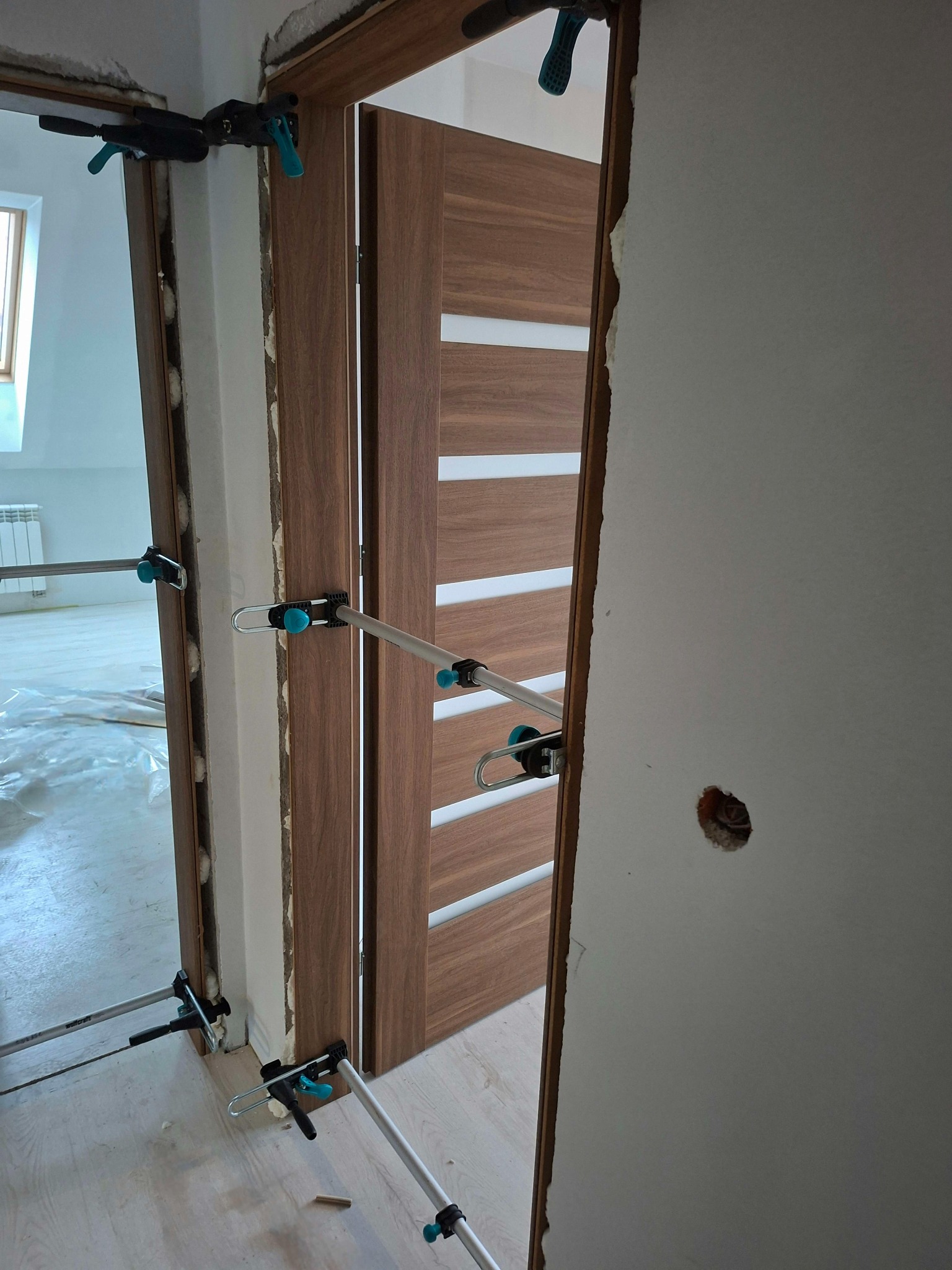 Montaż wewnętrznych drzwi z poziomymi przeszkleniami, przy użyciu rozpórek. Widoczna ościeżnica i fragment ściany z otworem na instalację elektryczną.