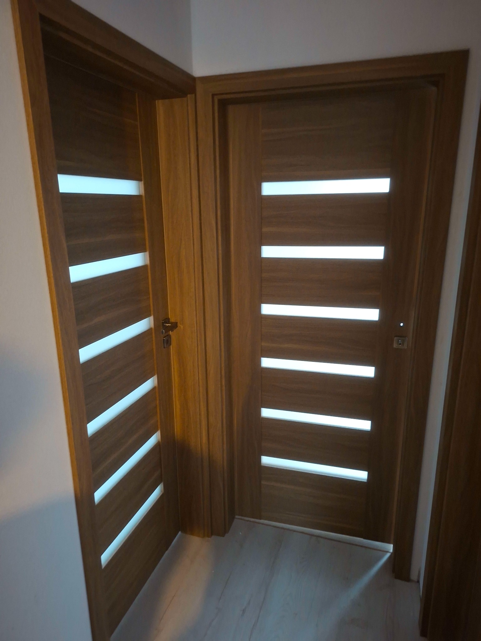 Dwa drewniane, nowoczesne drzwi wewnętrzne z poziomymi, białymi przeszkleniami, zamontowane w jasnym pomieszczeniu. Widoczne klamki i fragment podłogi.