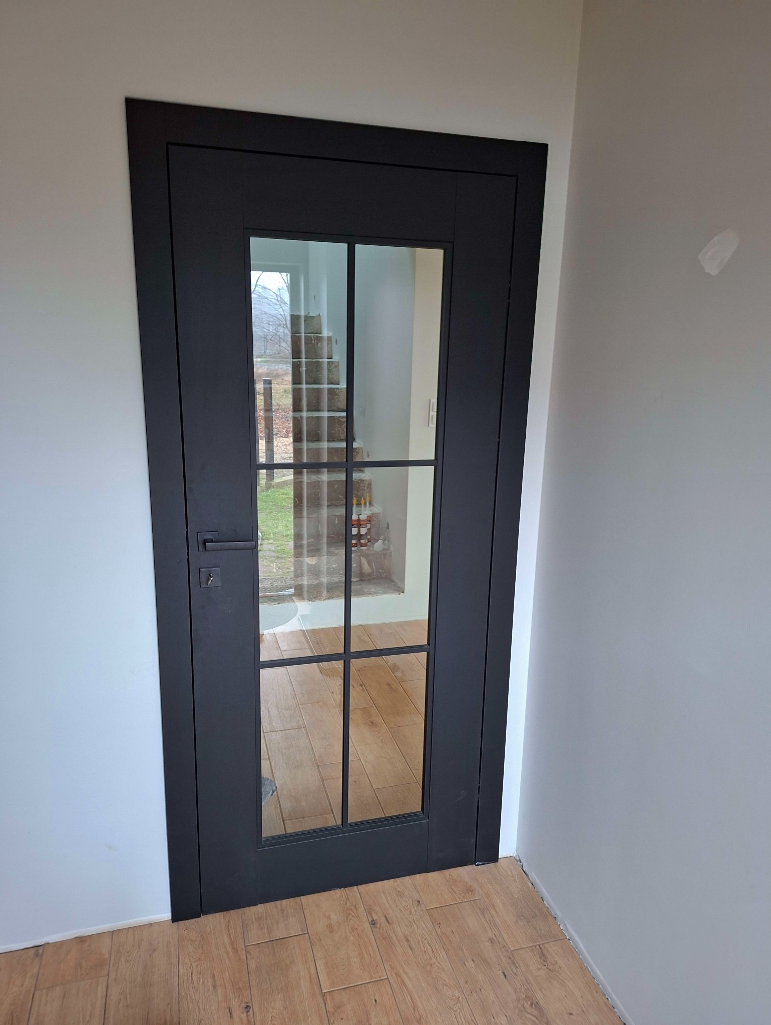 Czarne, przeszklone drzwi wewnętrzne z widokiem na schody, zamontowane w jasnym pomieszczeniu z drewnianą podłogą. Minimalistyczny design, nowoczesne wykończenie.