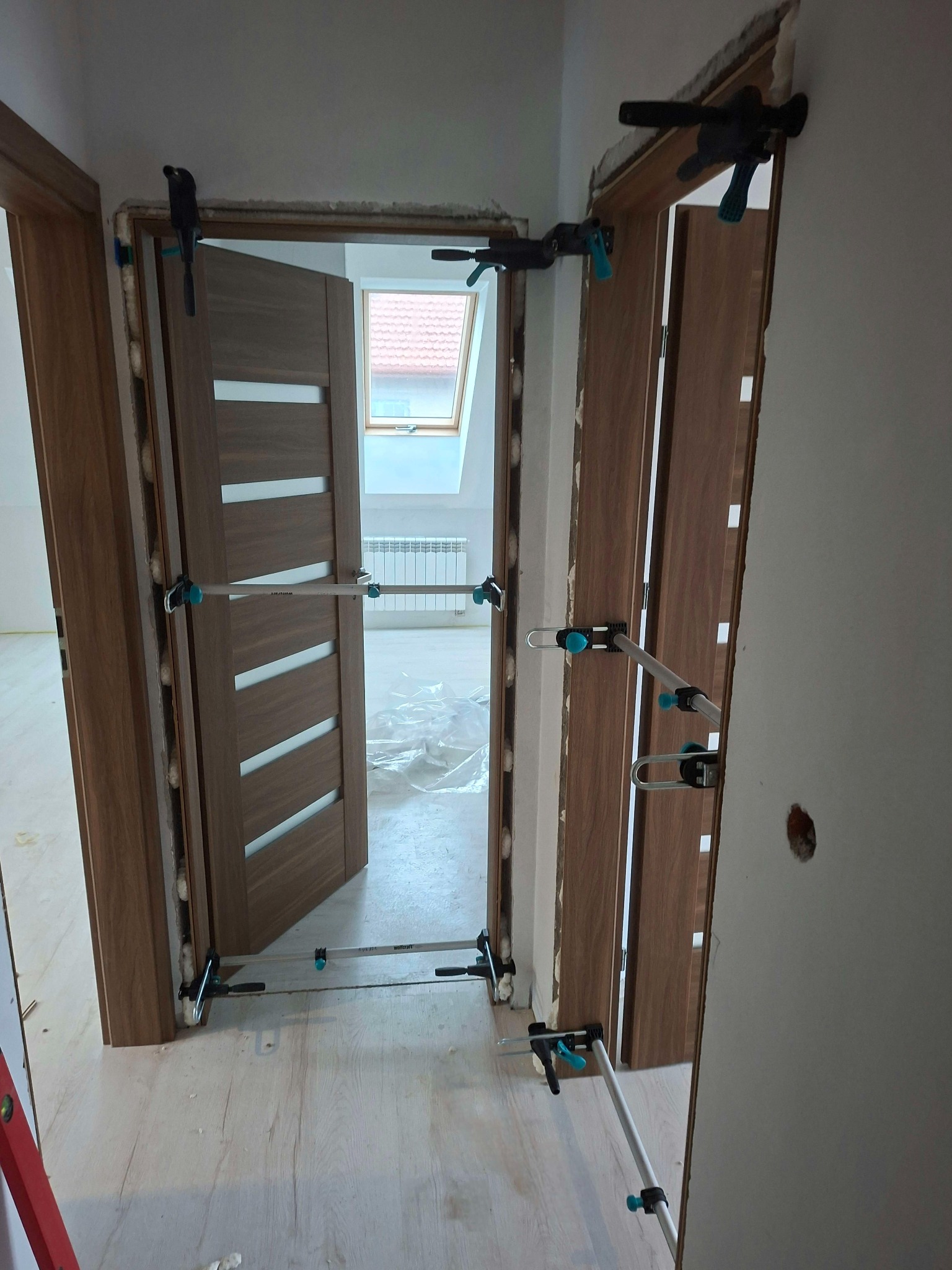 Montaż ościeżnicy drzwiowej z użyciem rozpór, widok na wnętrze z oknem dachowym. Drzwi z drewnopodobnym fornirem i białymi wstawkami, jasna podłoga.