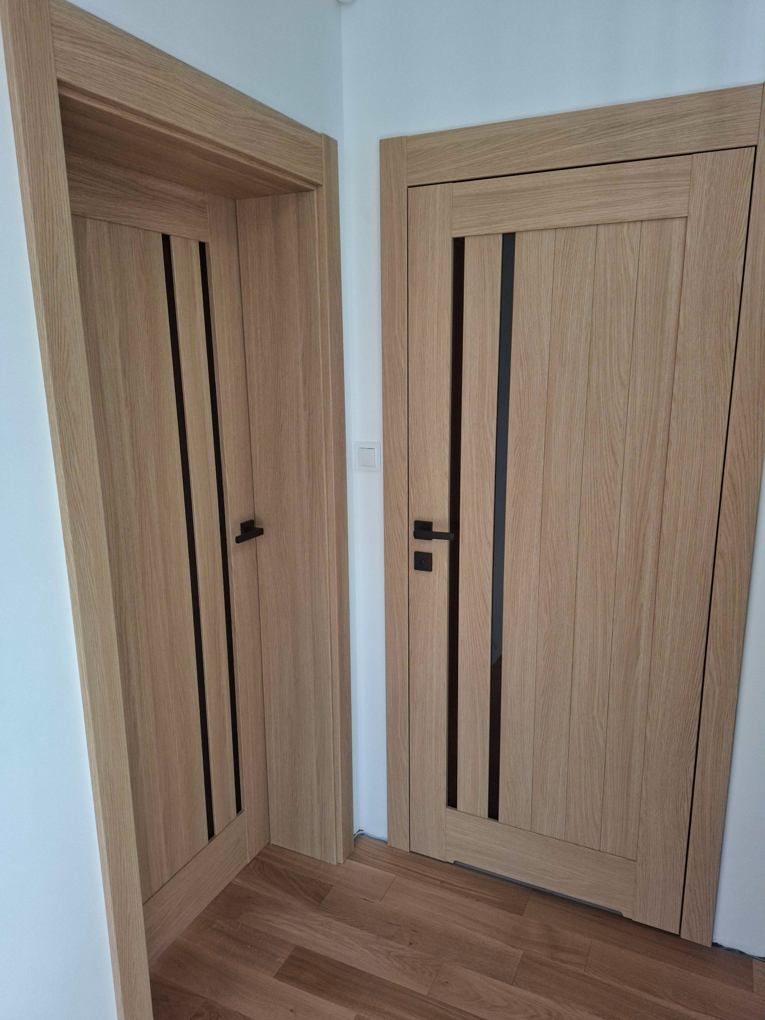 Dwa zamontowane, drewniane drzwi wewnętrzne z czarnymi akcentami i klamkami, widoczne na tle białej ściany i drewnianej podłogi.