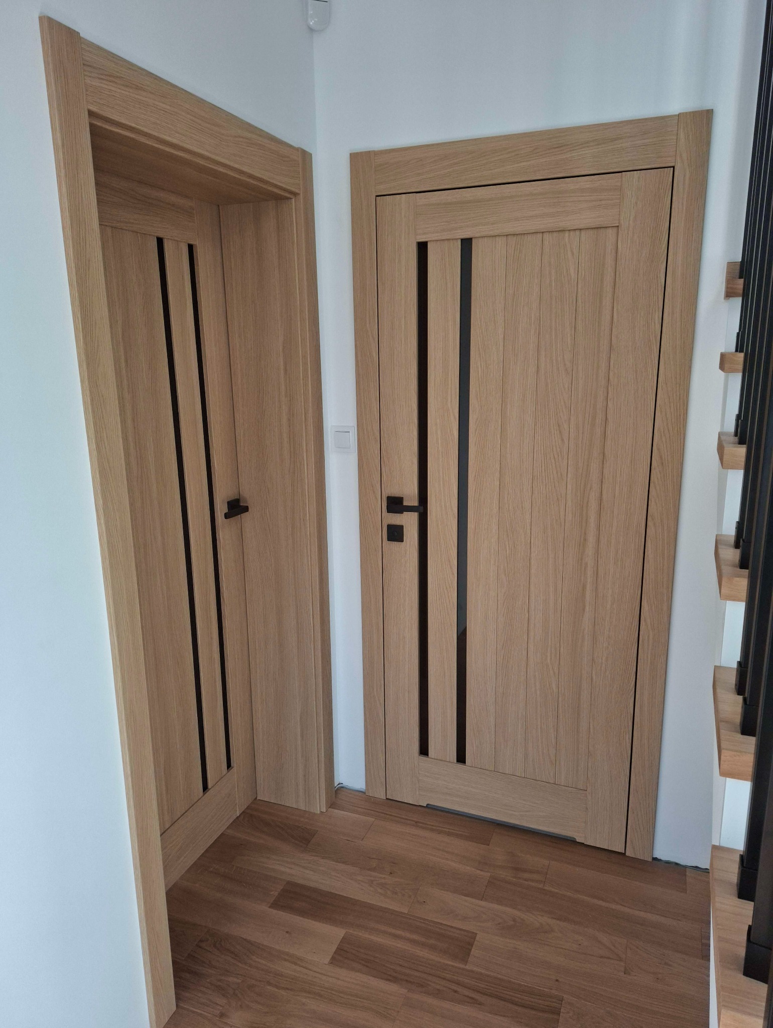 Dwie drewniane drzwi wewnętrzne z czarnymi akcentami, zamontowane w białym korytarzu z drewnianą podłogą i fragmentem schodów. Minimalistyczny design.