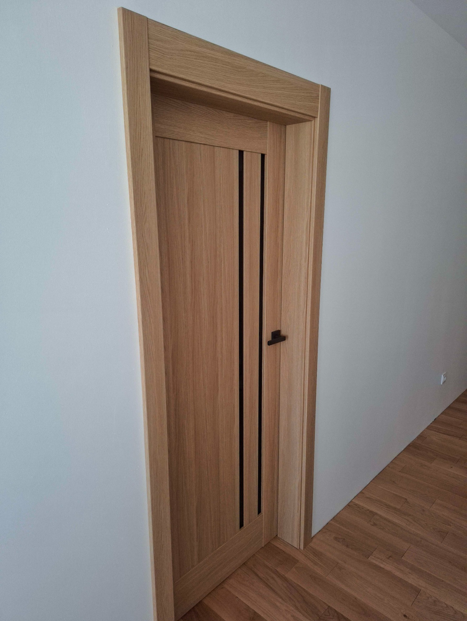 Zamontowane, drewniane drzwi wewnętrzne z czarnymi, pionowymi wstawkami i klamką na tle białej ściany i drewnianej podłogi. Widok z perspektywy.