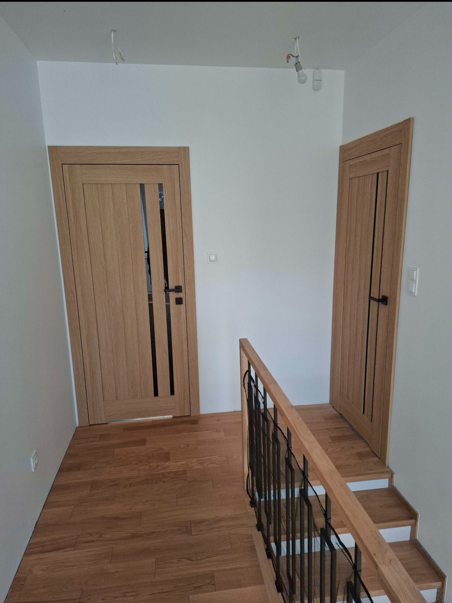 Jasne, drewniane drzwi wewnętrzne z przeszkleniem i czarną klamką, zamontowane w białym korytarzu z drewnianą podłogą i schodami. Widoczne niedokończone oświetlenie.