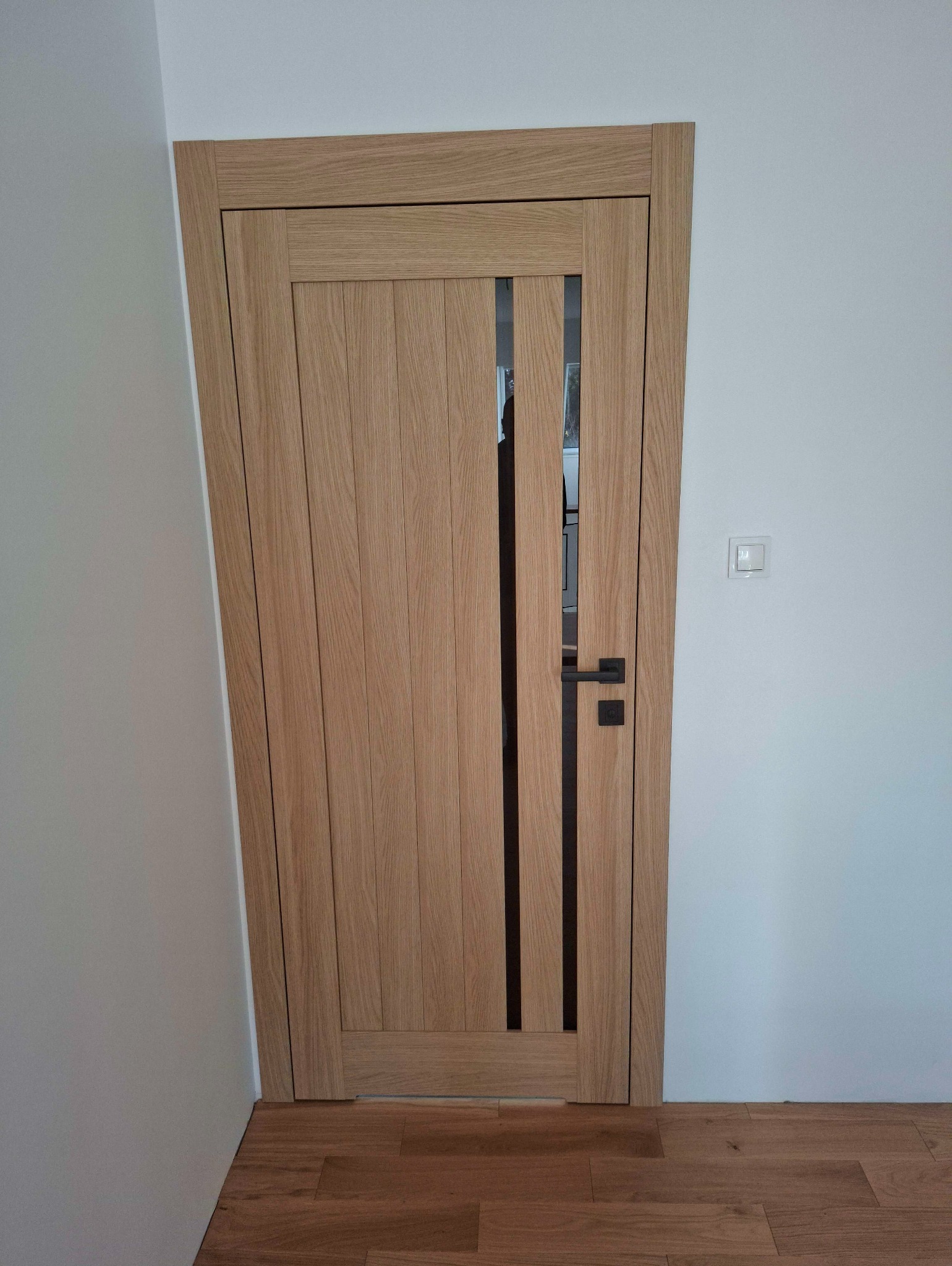 Zamontowane, dębowe drzwi wewnętrzne z czarnymi, wąskimi przeszkleniami i klamką, na tle białej ściany i drewnianej podłogi. Widoczny włącznik światła.