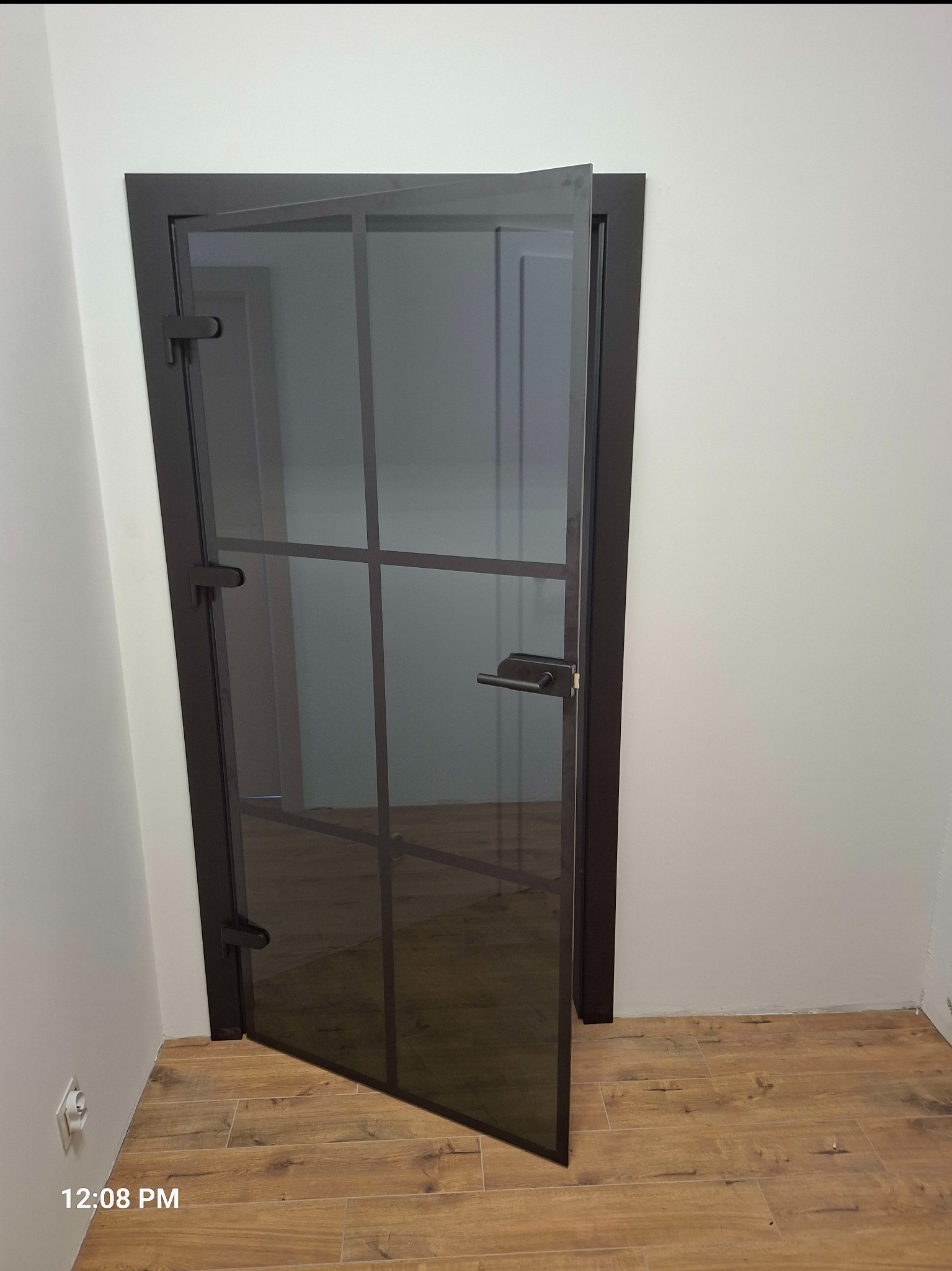 Ciemna, przeszklona, częściowo otwarta wewnętrzna rama drzwiowa z widoczną podłogą z paneli imitujących drewno i fragmentem białej ściany.