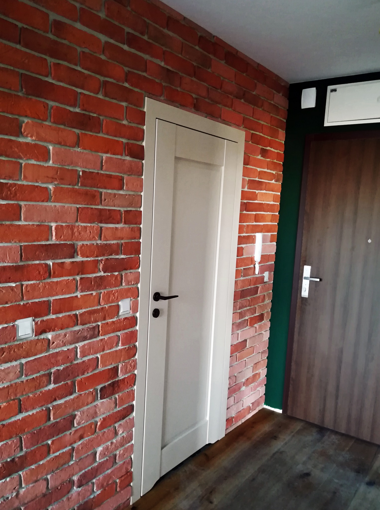 Ściana z czerwonej cegły z białymi drzwiami i drewnianą podłogą. Widoczny fragment brązowych drzwi wejściowych i zielony fragment ściany.