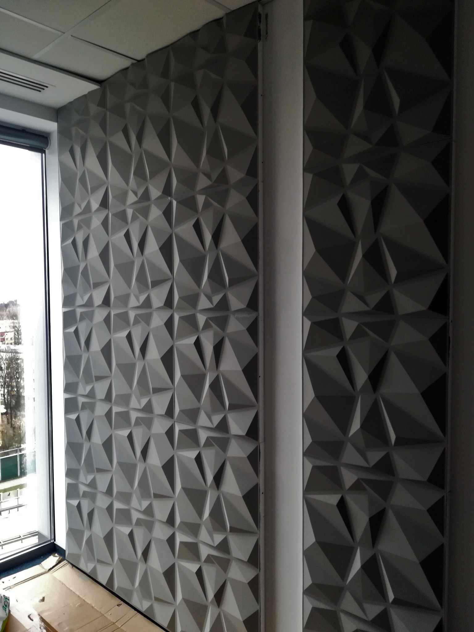 Panel ścienny 3D w szarym odcieniu, z geometrycznym wzorem trójkątów, zamontowany w nowoczesnym wnętrzu biurowym obok okna. Widoczne łączenia paneli.