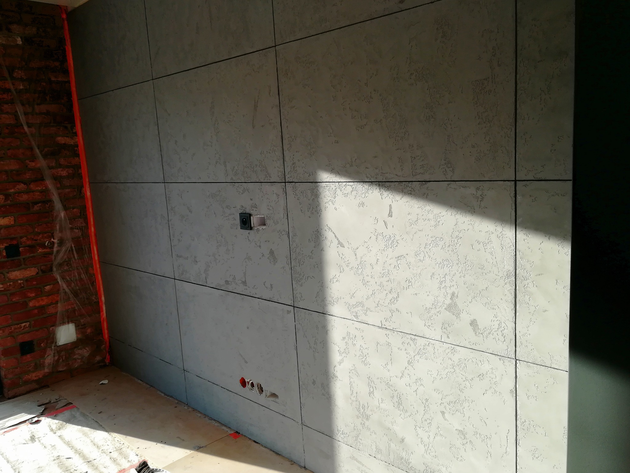 Ściana wykończona płytami imitującymi beton z widocznymi fugami, gniazdami elektrycznymi i fragmentem surowej cegły. W tle widoczne niedokończone prace.