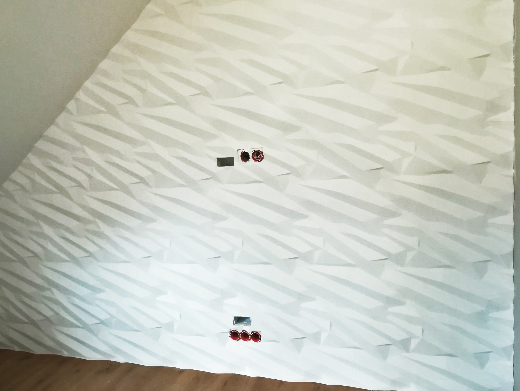 Ściana z dekoracyjną, trójwymiarową tapetą w odcieniach bieli. Widoczne gniazdka elektryczne i fragment podłogi. Ukośny sufit dodaje unikalności.