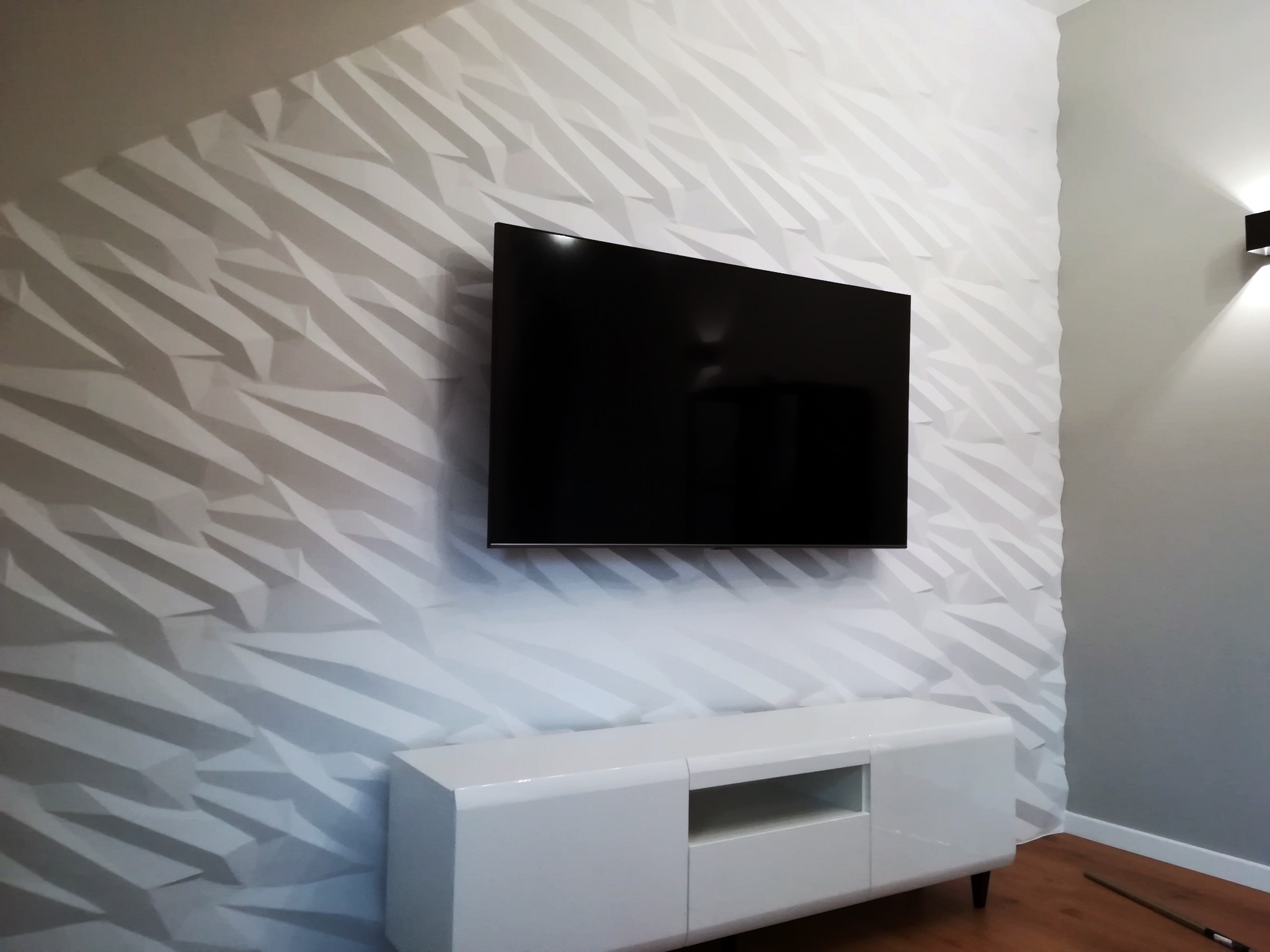 Nowoczesny salon z telewizorem na ścianie z dekoracyjną, trójwymiarową białą okładziną. Pod telewizorem biała szafka RTV. Minimalistyczny design.