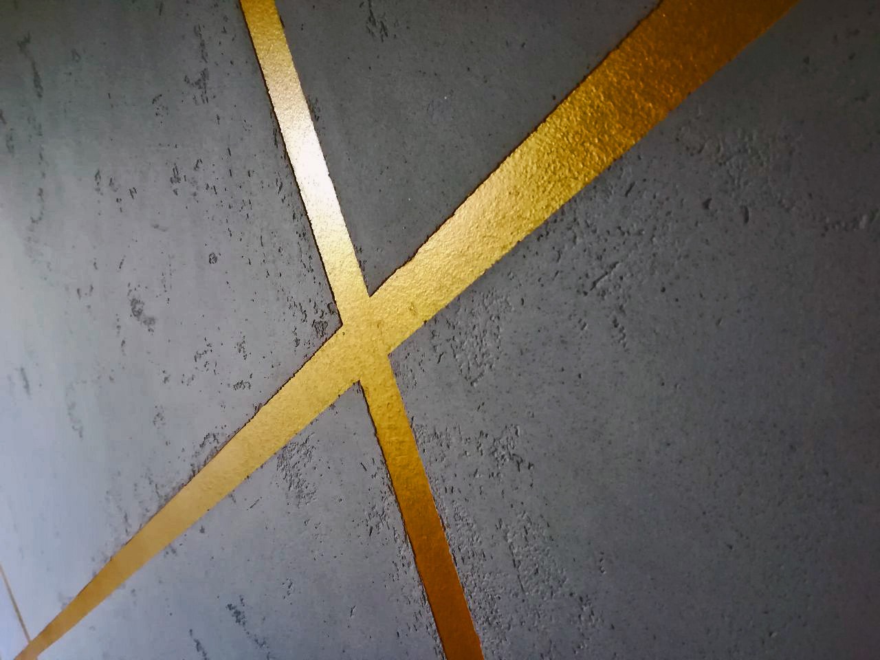 Szara ściana z dekoracyjnymi, złotymi pasami tworzącymi geometryczny wzór. Efektowna faktura i połyskujące złoto dodają wnętrzu nowoczesnego charakteru.