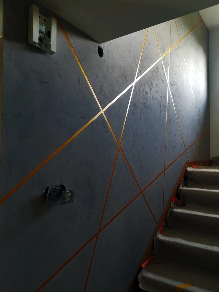 Szara ściana z dekoracyjnymi, złotymi liniami tworzącymi geometryczny wzór. Widoczne schody i gniazdka elektryczne. Nowoczesne wykończenie wnętrza.