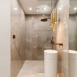 STARMACH STUDIO KAROLINA SMOTER-STARMACH - Elegancka łazienka z kabiną prysznicową typu walk-in, umywalką cylindryczną i złotą armaturą. Beżowe płytki imitujące marmur, oświetlenie punktowe i lustro.