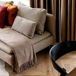 STARMACH STUDIO KAROLINA SMOTER-STARMACH - Beżowa sofa z dekoracyjnymi poduszkami w odcieniach czerwieni i brązu, pled z frędzlami, dywan w krowie łaty i fragment krzesła na drewnianej podłodze. Stonowana kolorystyka.