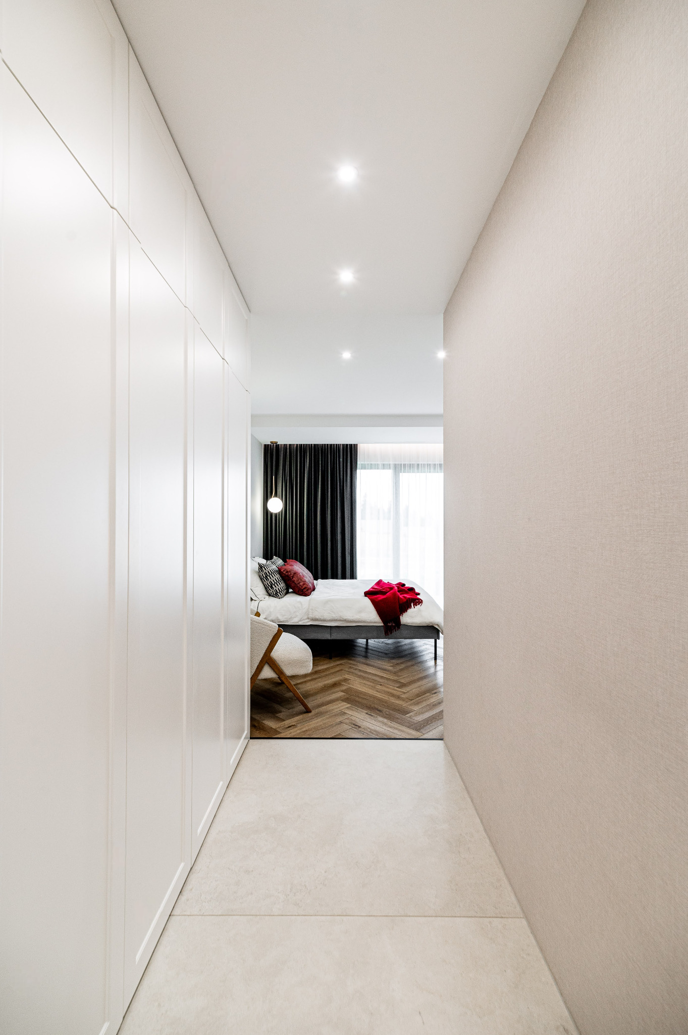 Nowoczesna sypialnia z garderobą w minimalistycznym stylu. Parkiet w jodełkę, białe ściany i szafy w zabudowie. Akcent kolorystyczny: czerwona narzuta na łóżku.