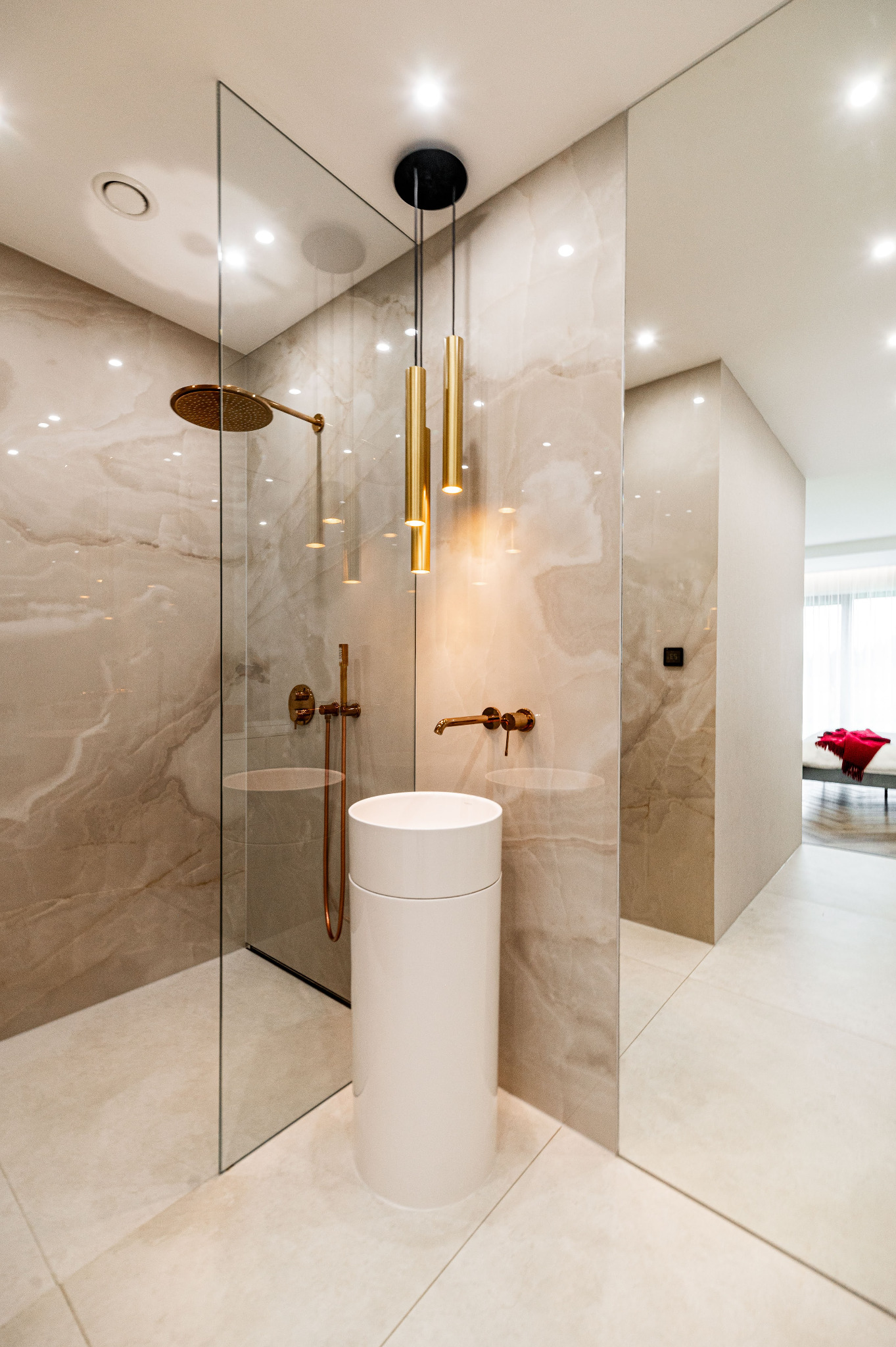 Elegancka łazienka z kabiną prysznicową typu walk-in, umywalką kolumnową i złotą armaturą. Beżowe płytki imitujące marmur i designerska lampa wisząca.
