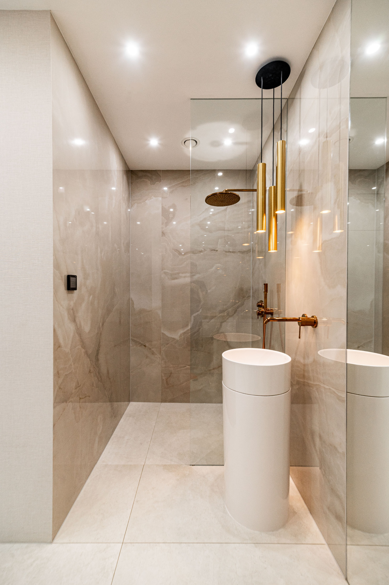 Elegancka łazienka z kabiną prysznicową typu walk-in, umywalką cylindryczną i złotą armaturą. Beżowe płytki imitujące marmur, oświetlenie punktowe i lustro.
