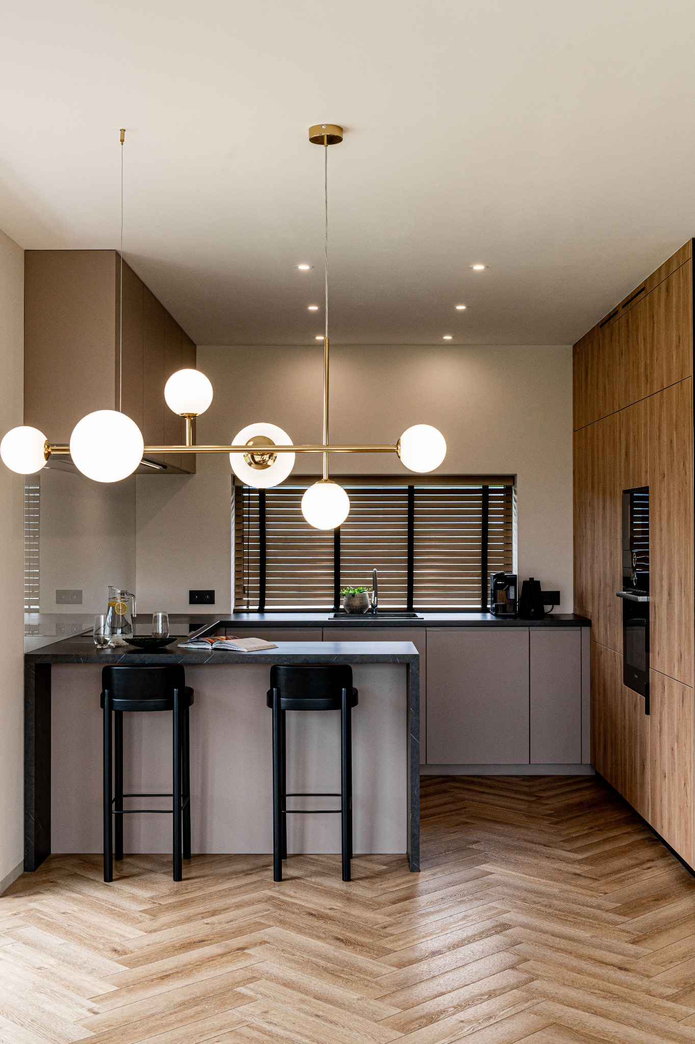 Nowoczesna kuchnia z wyspą, drewnianą podłogą w jodełkę i złotą lampą wiszącą z kulistymi kloszami. Minimalistyczne krzesła barowe i zabudowa kuchenna w odcieniach beżu i drewna.