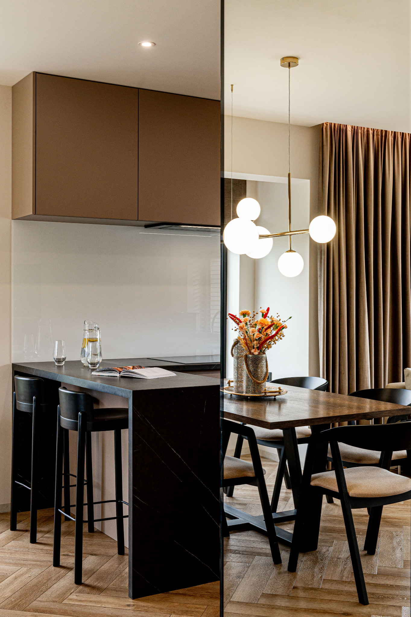 Nowoczesna kuchnia z wyspą i jadalnią, drewniana podłoga w jodełkę, czarne krzesła barowe i stół, złoty żyrandol, beżowe zasłony, wazon z suszonymi kwiatami.