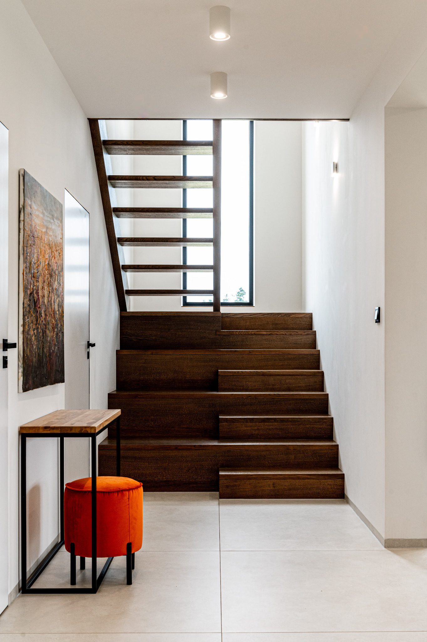 Nowoczesne schody drewniane w jasnym holu, z pomarańczowym pufem i abstrakcyjnym obrazem. Minimalistyczne oświetlenie punktowe.