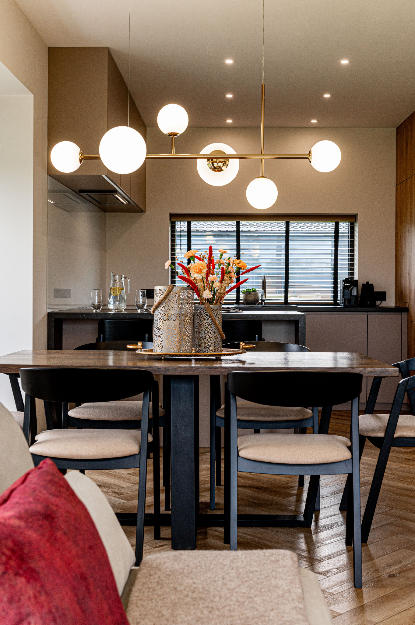 Jadalnia z drewnianym stołem, czarnymi krzesłami i dekoracyjnym bukietem. Nad stołem nowoczesna lampa z kulistymi kloszami. Parkiet w jodełkę na podłodze.